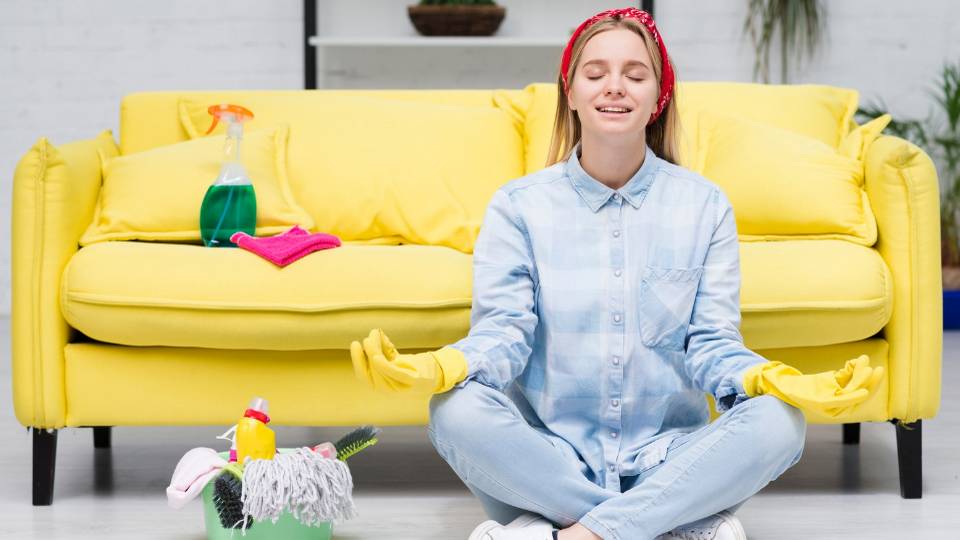 mulher sentada no chão em frente ao sofá, usando luvas de borracha de limpeza, em posição de lótus, como se estivesse meditando