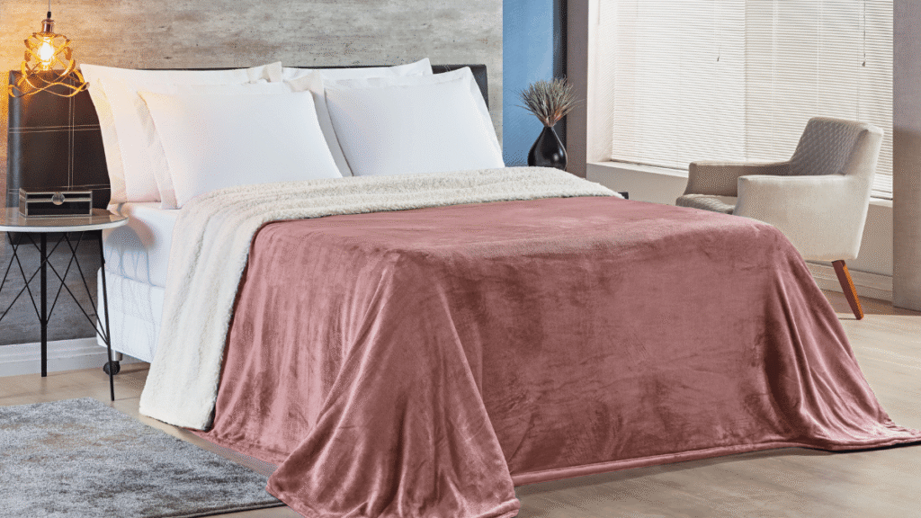 Cobertor Alaska rosa envelhecido com forro de sherpa branco estendido em cama de casal, em ambiente com poltrona e luminária moderna.