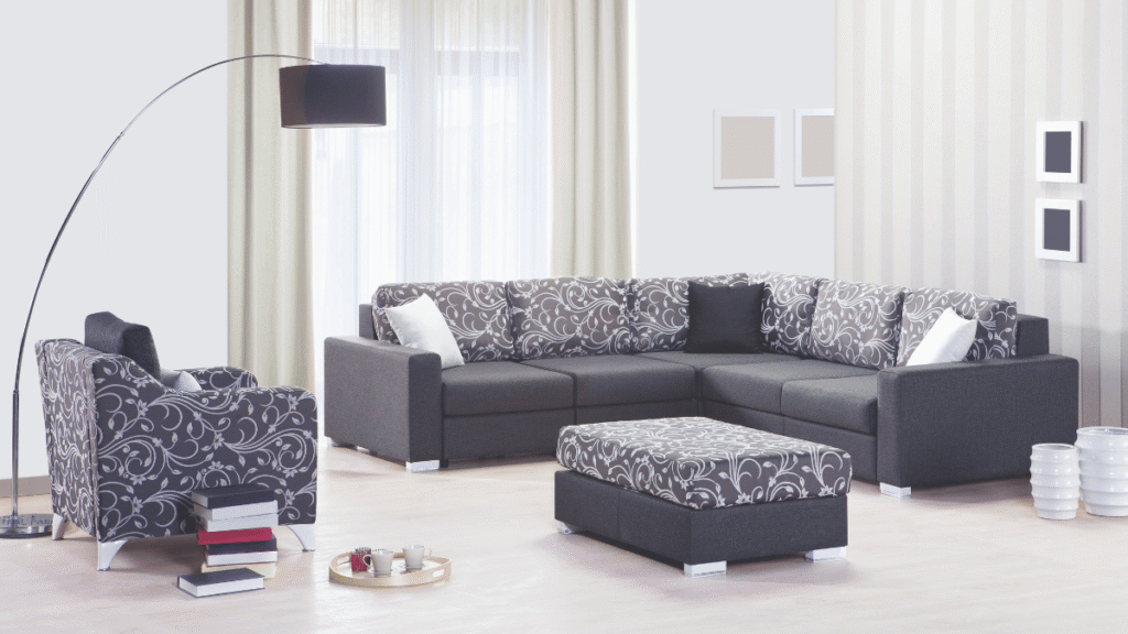 Sala moderna com sofá de canto cinza-escuro, almofadas e estofado com capa estampada em arabescos brancos, poltrona combinando, puff central e decoração minimalista em tons neutros.