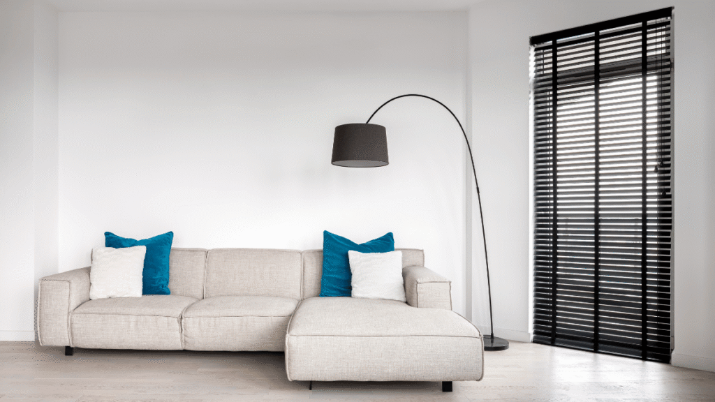 Sala minimalista com sofá de canto bege claro, almofadas brancas e azuis, luminária de chão preta e persiana escura em janela ampla, em um ambiente moderno e clean.