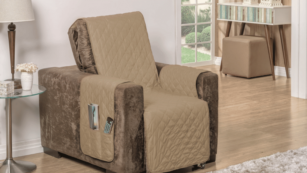 Poltrona marrom com capa protetora bege acolchoada, com bolsos laterais contendo controle remoto e revista, em uma sala com piso de madeira clara, mesa lateral de vidro com abajur e vaso de flores, além de um aparador com livros e pufe ao fundo, próximo a uma grande janela com vista para um jardim.