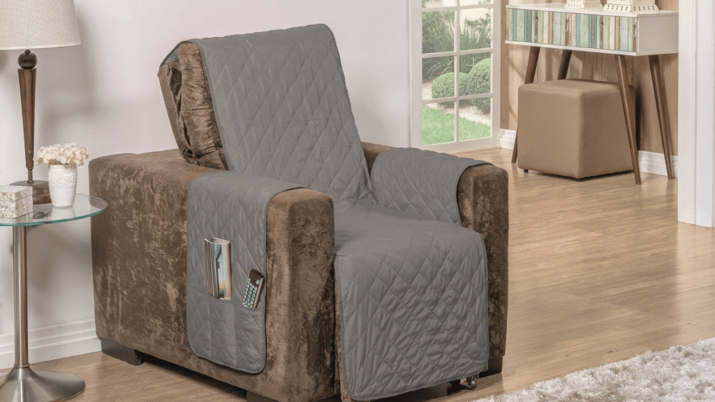 Poltrona marrom com capa protetora cinza acolchoada, com bolsos laterais contendo controle remoto e revista, posicionada em uma sala com piso de madeira clara, mesa lateral de vidro com abajur e flores, além de uma bancada com livros e pufe ao fundo, próximo a uma grande janela com vista para um jardim.