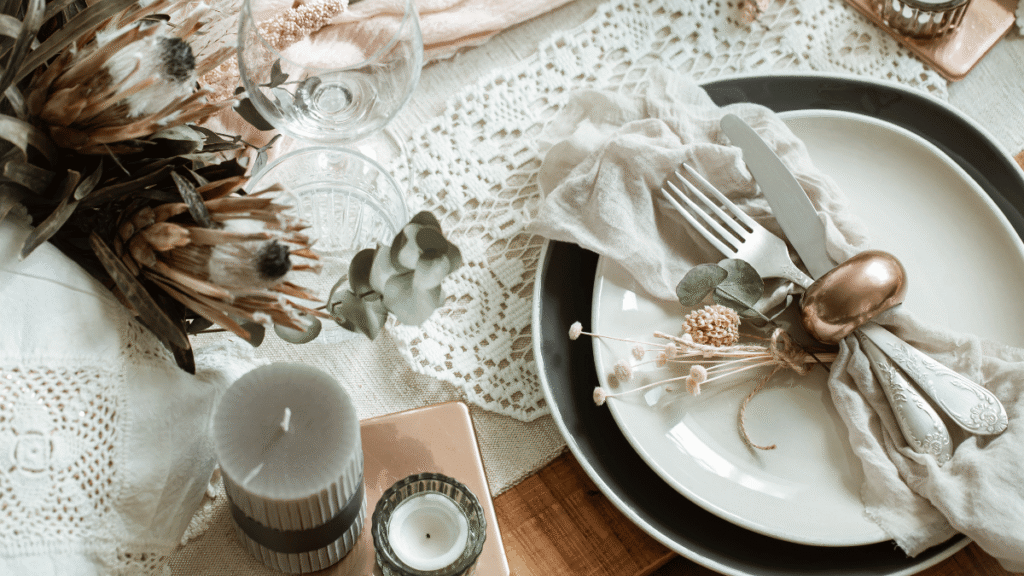 Mesa posta em estilo rústico e elegante, com pratos em tons neutros sobre uma toalha de crochê, talheres com acabamento vintage dispostos sobre um guardanapo de tecido, detalhes de folhagens secas e flores desidratadas, além de velas decorativas ao lado de um arranjo de flores em tons terrosos.