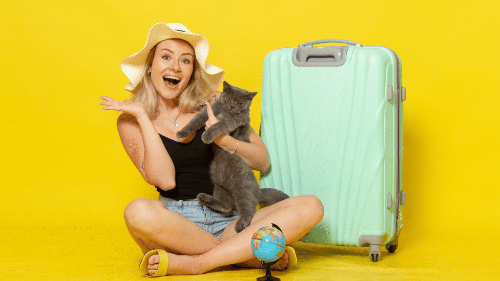 Mulher sorridente sentada no chão com um gato cinza no colo, ao lado de uma mala de viagem verde-clara e um globo terrestre pequeno, em frente a um fundo amarelo vibrante.