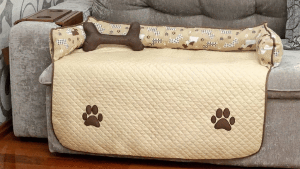 Protetor acolchoado bege com bordas marrons e duas patinhas estampadas, posicionado sobre o assento de um sofá cinza, acompanhado de almofadas laterais com estampa de cachorrinhos e uma almofada central em formato de osso marrom, ideal para pets.