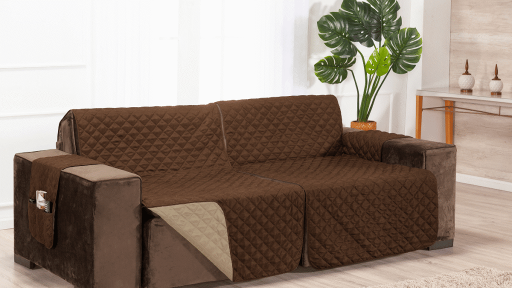 Sofá marrom com protetor acolchoado dupla face em tom chocolate, cobrindo assento, encosto e braços, com bolso lateral e ambiente decorado com planta verde e elementos em madeira clara.