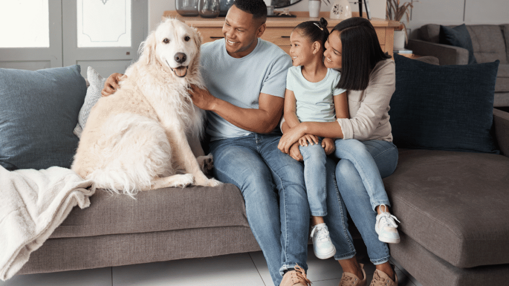 Família sorridente sentada em um sofá cinza ao lado de um cachorro grande e peludo de cor clara, dentro de uma sala de estar aconchegante, com almofadas e manta decorativa.